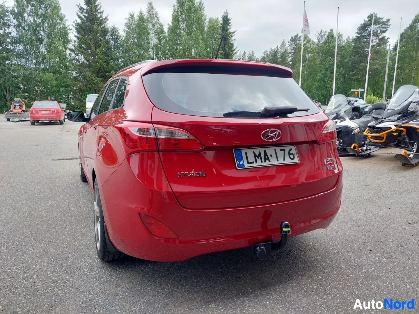 Takaa: Punainen Hyundai i30 farmari takaa kuvattuna. Autossa on vetokoukku, takalasinpyyhin ja rekisterikilpi. Takana näkyy moottorikelkkoja