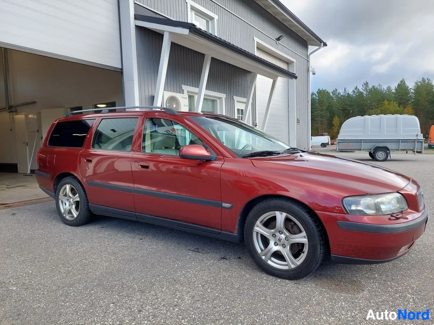 Etuviisto, nokka oikealle: Punainen henkilöauto seisoo ulkona rakennuksen edessä. Auto on kuvattu hieman edestä ja sivulta vasemmalta puolelta. Siinä on kattokiskot ja kevytmetalli