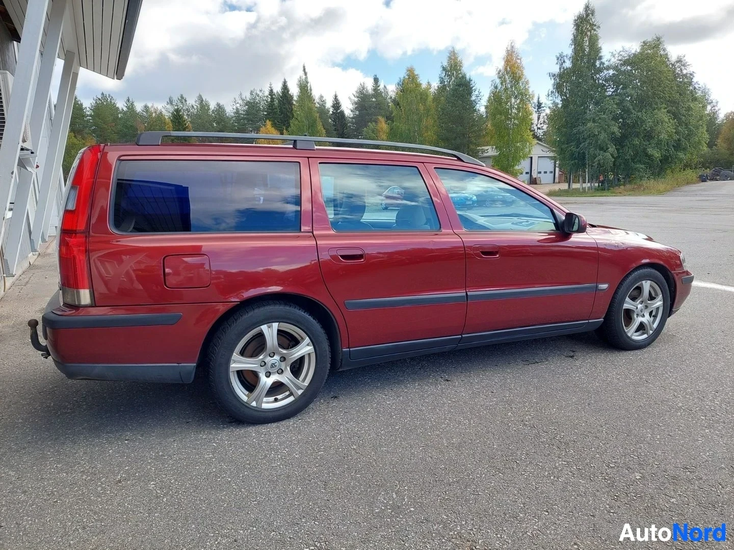 Sivulta: Punainen henkilöauto on kuvattu ulkona oikealta sivulta. Siinä on kattokiskot, kevytmetallivanteet ja vetokoukku.