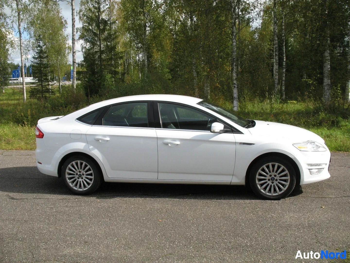 Sivulta: Valkoinen Ford Mondeo henkilöauto kuvattuna suoraan sivulta, pysäköitynä päällystetylle tielle puurivin eteen.
