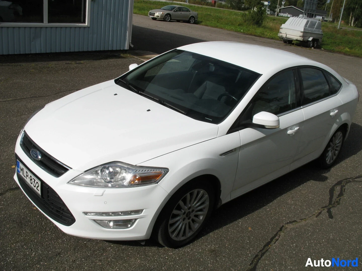 Etuviisto, nokka oikealle: Valkoinen Ford Mondeo henkilöauto kuvattuna yläviistosta edestä ja vasemmalta. Auto on pysäköity asfaltille rakennuksen ja taustalla näkyvän peräkärrylli