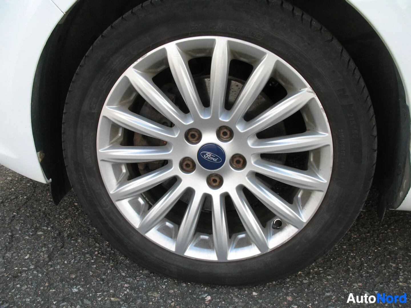 Yksityiskohta: Lähikuva auton etupyörästä, jossa on hopeinen monipuolainen kevytmetallivanne ja musta rengas. Rengas on hieman kulunut. Vanteen keskellä on sininen Ford-logo.