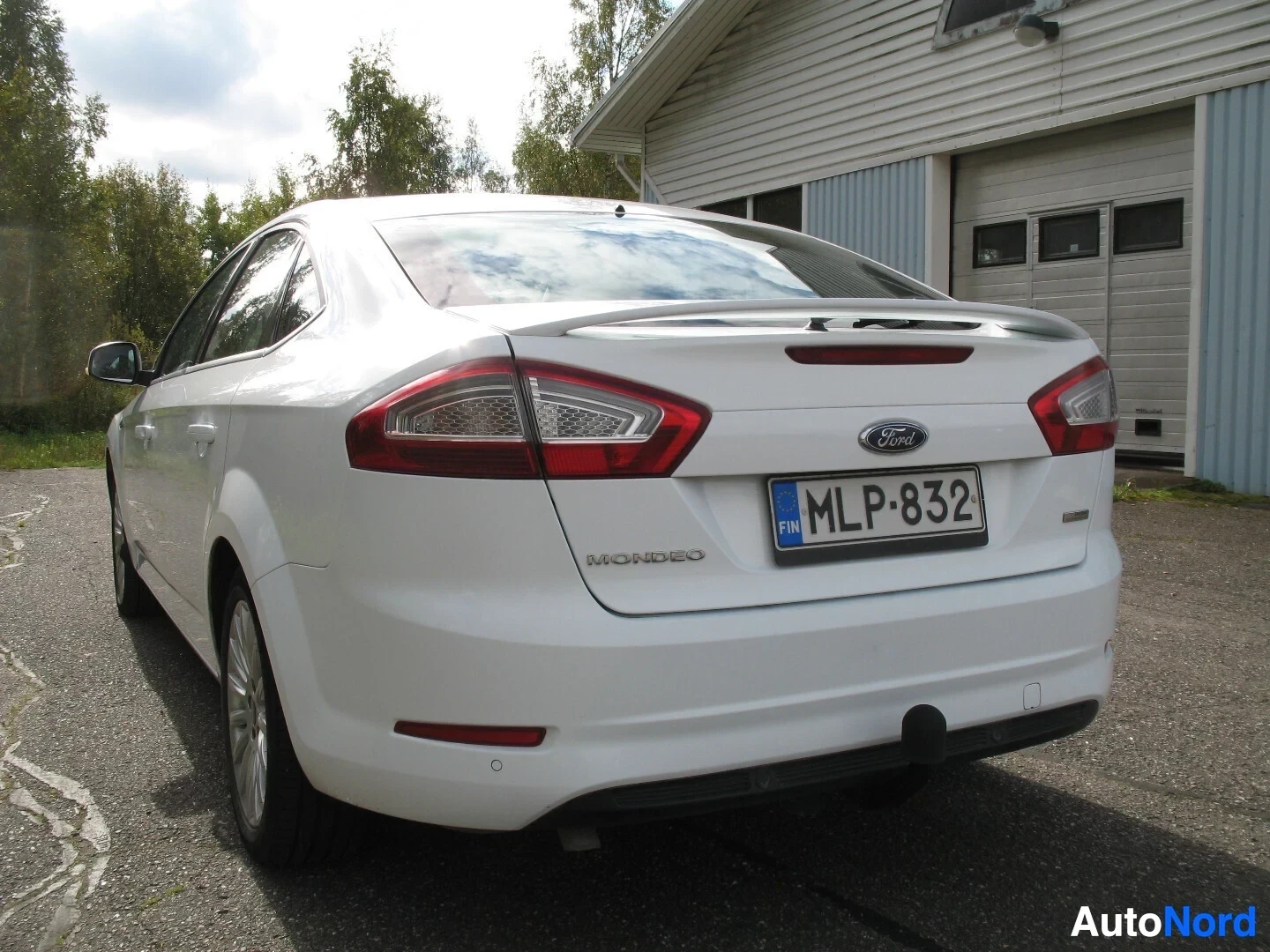 Takaviisto: Valkoinen Ford Mondeo -henkilöauto, takapää ja vasen kylki näkyvissä, takana rakennus ja puita.