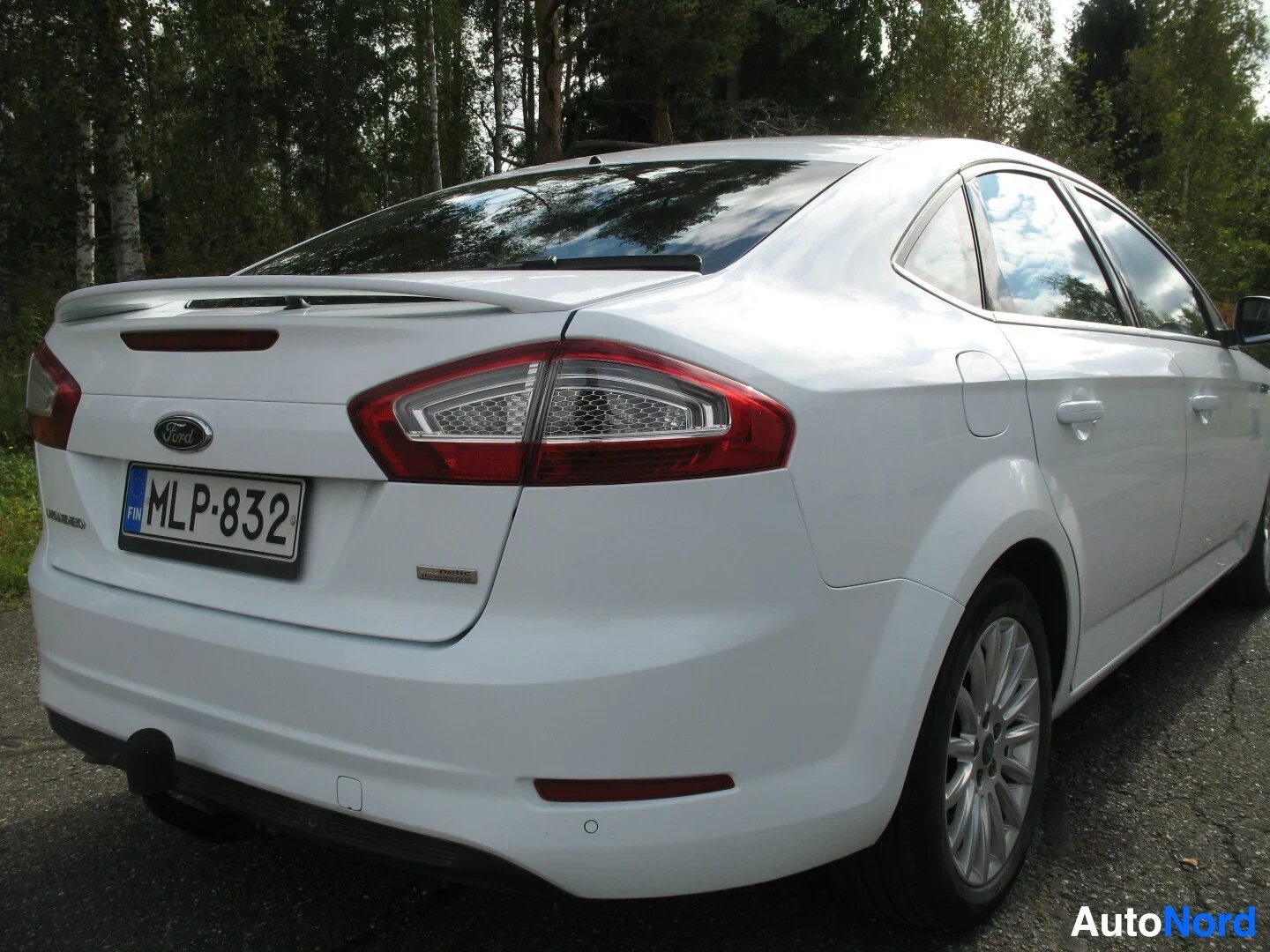 Takaviisto: Valkoisen Ford Mondeo henkilöauton oikea takakulma, ajoneuvo on pysäköity ulos, taustalla puita.