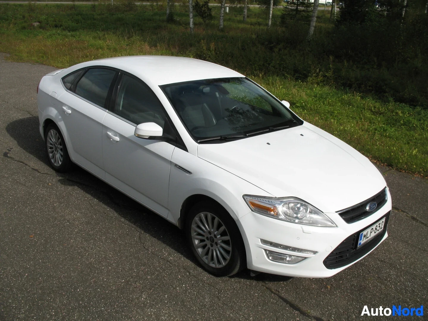 Etuviisto, nokka oikealle: Valkoinen Ford Mondeo henkilöauto kuvattuna edestä ja vasemmalta, pysäköitynä päällystetylle tielle metsäisen taustan edessä.