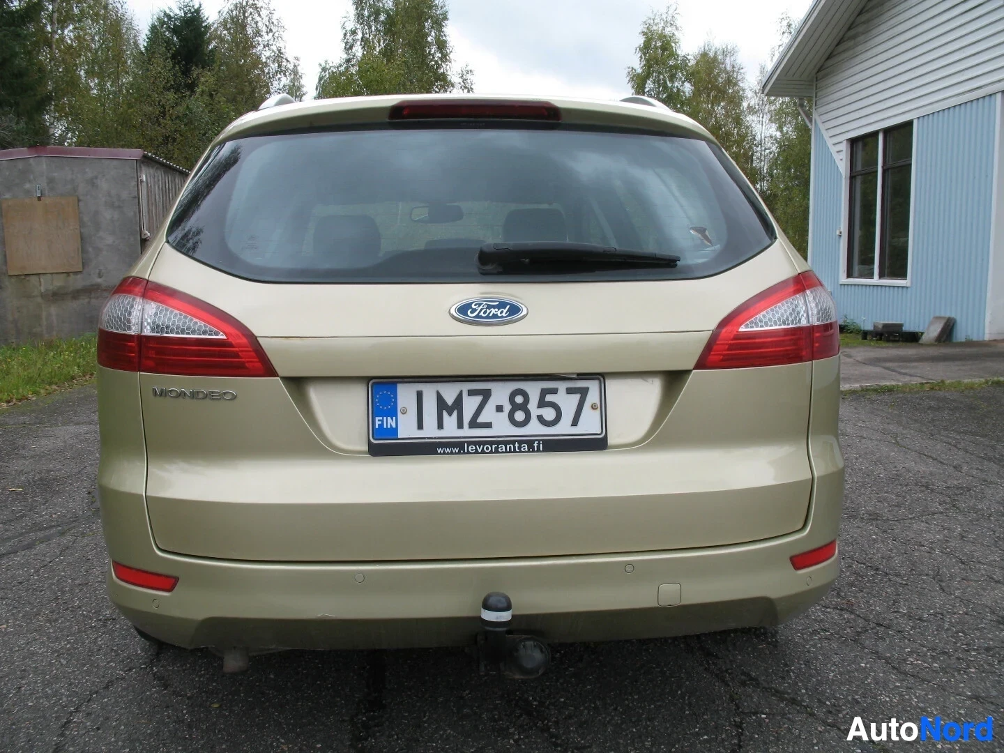Takaa: Kultaisen farmariauton takaosa suoraan edestä, jossa näkyy peräkontin luukku, takavalot, Ford-logo, rekisterikilpi "" ja vetokoukku. Rekisterikilven alla