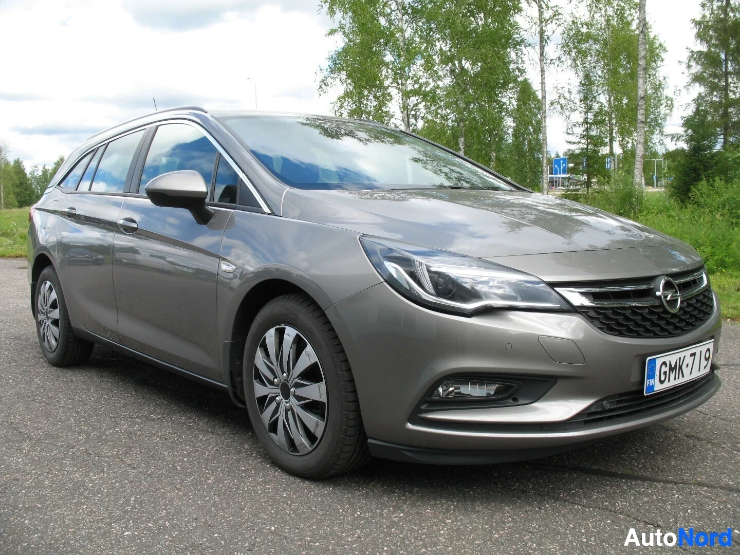 Etuviisto, nokka oikealle: Harmaa Opel Astra Sports Tourer on kuvattu etuoikealta ulkona kirkkaassa säässä. Autossa on mustat alumiinivanteet ja kattoikkuna. Ajovalot ovat päällä.