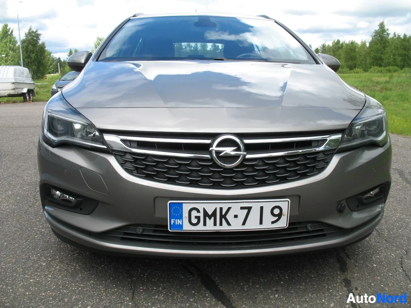 Edestä: Harmaan Opel Astra Sports Tourerin etuosa on kuvattu suoraan edestä ulkona kirkkaassa säässä. Rekisterinumero on. Edessä näkyy pysäköintitunnistimet