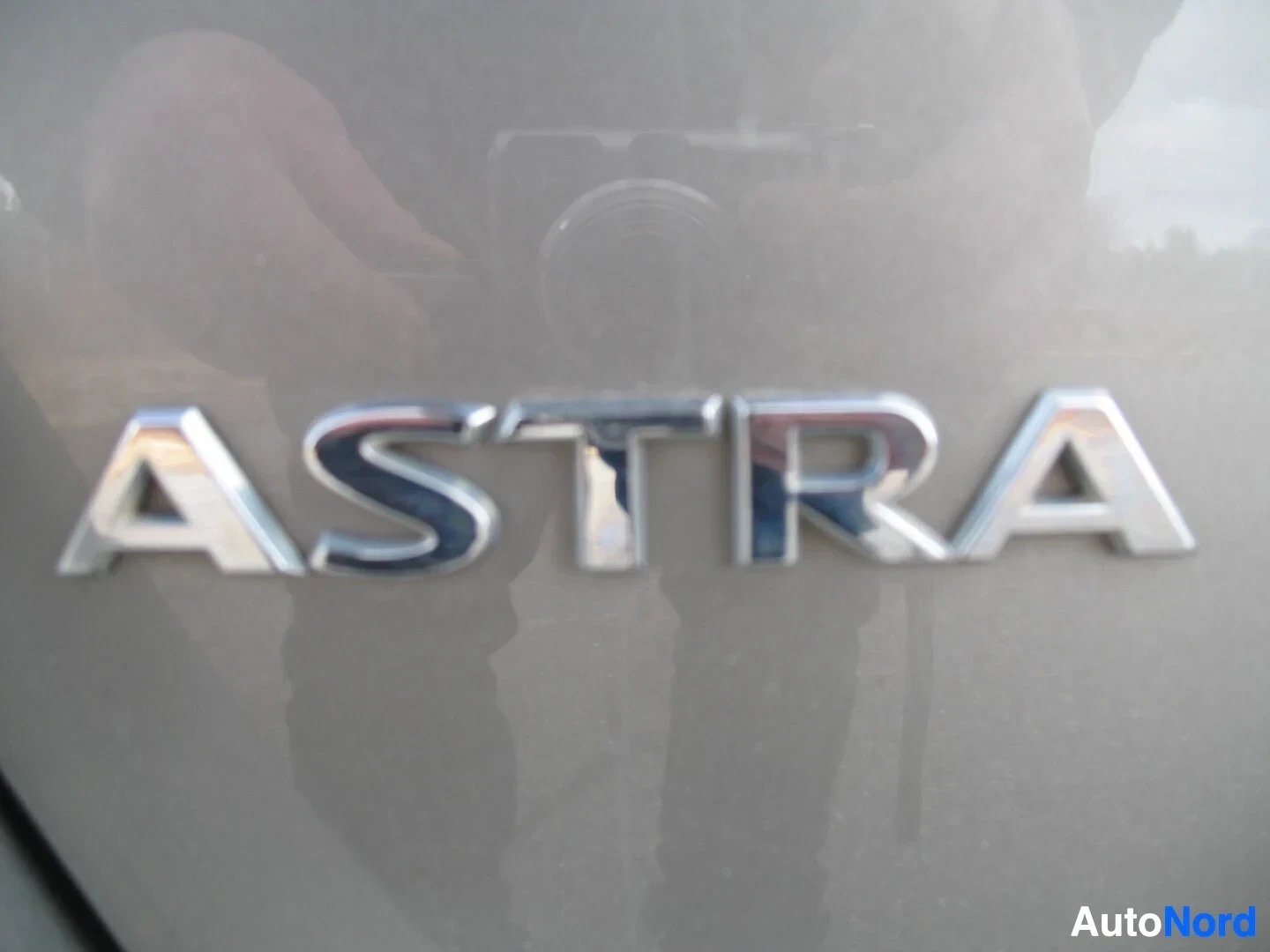 Yksityiskohta: Lähikuva auton kyljessä olevasta kromisesta "ASTRA"-merkistä, jossa näkyy heijastuksia kamerasta ja henkilöstä.