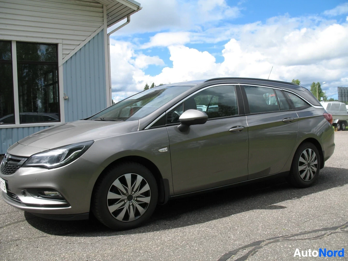 Sivulta: Harmaa Opel Astra Sports Tourer on kuvattu sivulta ulkona kirkkaassa säässä. Autossa on mustat alumiinivanteet ja kattokiskot. Takana näkyy auton perävaunun vet