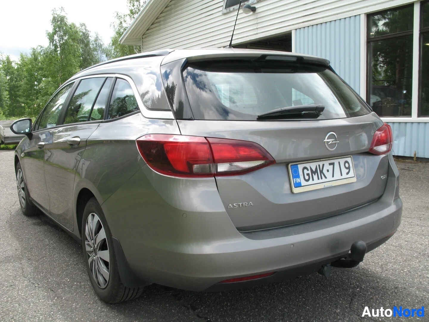 Takaviisto: Takaa oikealta puolelta otettu kuva harmaasta farmariautosta, joka on pysäköity rakennuksen eteen. Autossa on kattokiskot ja kevytmetallivanteet, ja takavalot o
