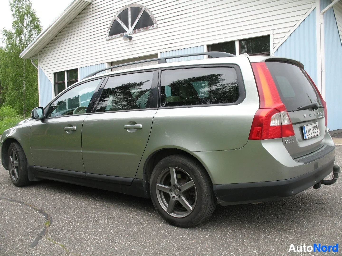 Takaviisto: Vaaleanvihreä Volvo V70 -farmariauto parkkipaikalla, kuvattuna takaa ja vasemmalta sivulta. Autossa on kattokaiteet, viisipuolaiset alumiinivanteet ja vetokoukk