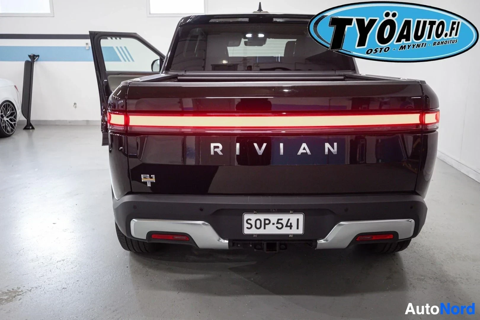 Takaa: Musta avolava-auto on pysäköity sisätilaan. Auton takaosa on näkyvissä, takaluukku on auki. Takaluukussa lukee "RIVIAN" ja sen yläpuolella on punainen vaakasuor