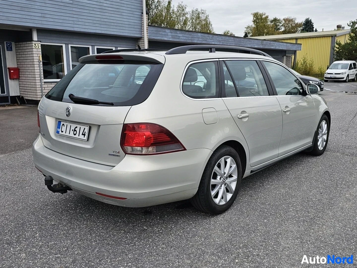 Takaviisto: Hopeanvärinen Volkswagen Golf Variant, jossa on kattokaiteet, alumiinivanteet ja vetokoukku, pysäköitynä rakennuksen eteen. Auton takaosa ja oikea kylki ovat nä