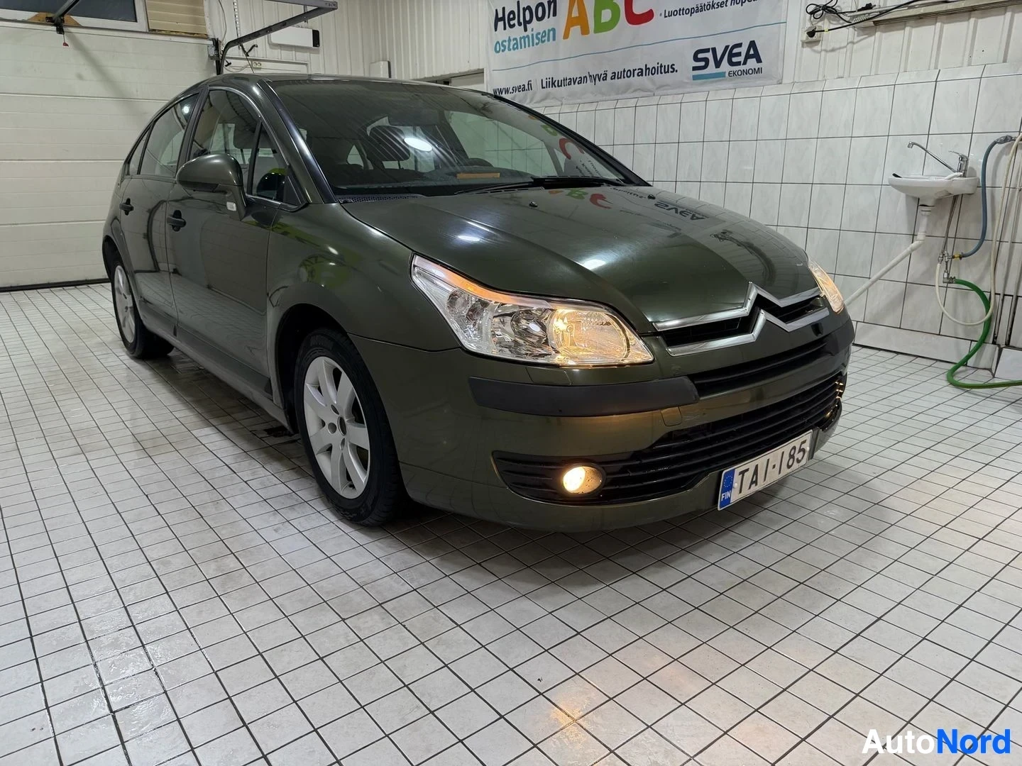 Etuviisto, nokka oikealle: Tummanvihreä Citroën C4 henkilöauto pysäköitynä valkoisilla ruutulaatoilla päällystetyn hallin sisällä. Auton etuosa ja vasen kylki näkyvät. Edessä rekis