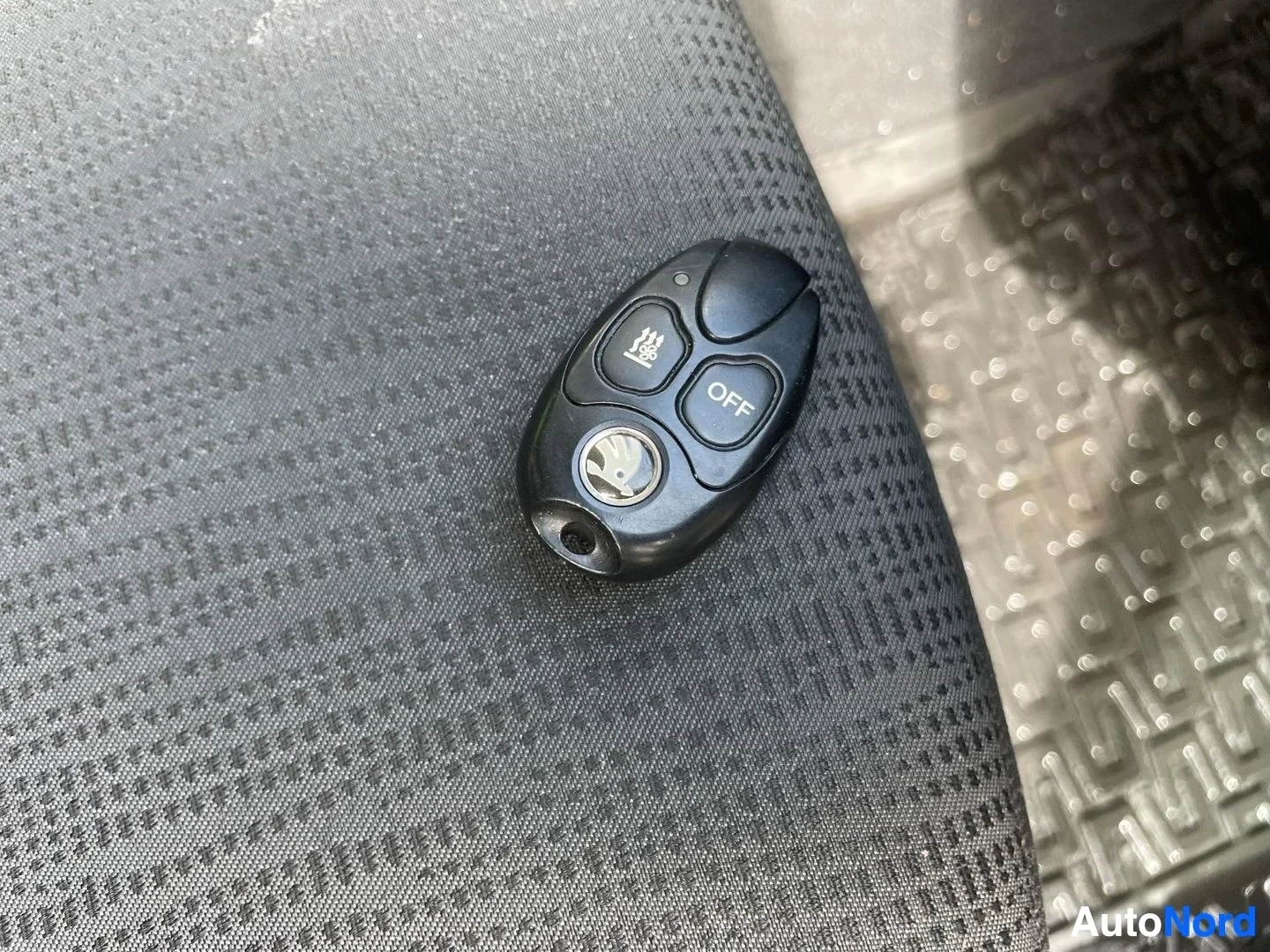 Yksityiskohta: Musta avaimenperä, jossa on Škoda-logo ja kolme painiketta, mukaan lukien OFF-merkitty painike, makaa tummalla kangasistuimella.