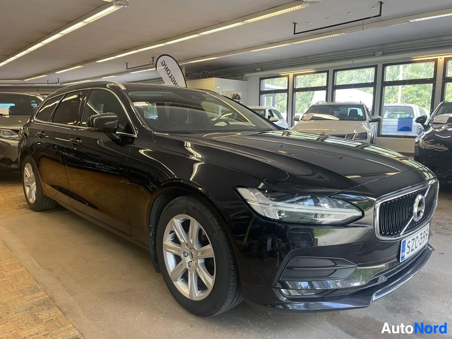 Etuviisto, nokka oikealle: Musta Volvo V90 -farmariauto kuvattuna edestä ja oikealta sivulta sisätiloissa. Auton rekisterikilpi on. Valot heijastuvat auton mustasta maalipinnasta.