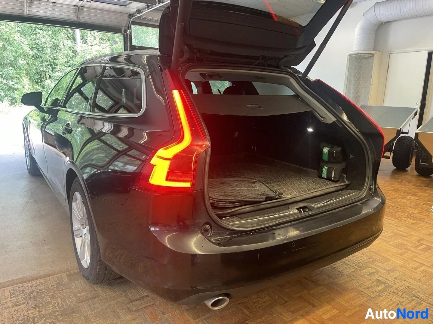 Tavaratila: Mustan Volvo V90 -farmariauton takaluukku on auki ja tavaratila näkyy. Tavaratilassa on kumimatto ja muutama pakkaus. Takavalot palavat.