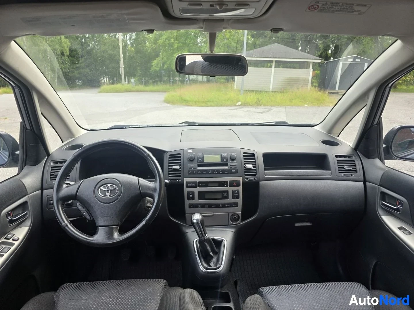 Muu: Näkymä ohjaamosta tuulilasin läpi: Toyota-merkkinen ohjauspyörä, keskikojelaudan radio ja ilmastointilaitteet, manuaalivaihteen keilu.