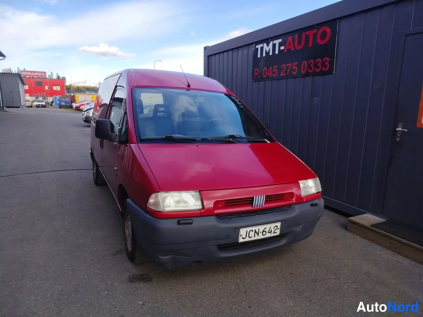Etuviisto, nokka oikealle: Punainen Fiat Scudo -pakettiauto etuviistosta oikealta, TMT-Auto-kontti taustalla.