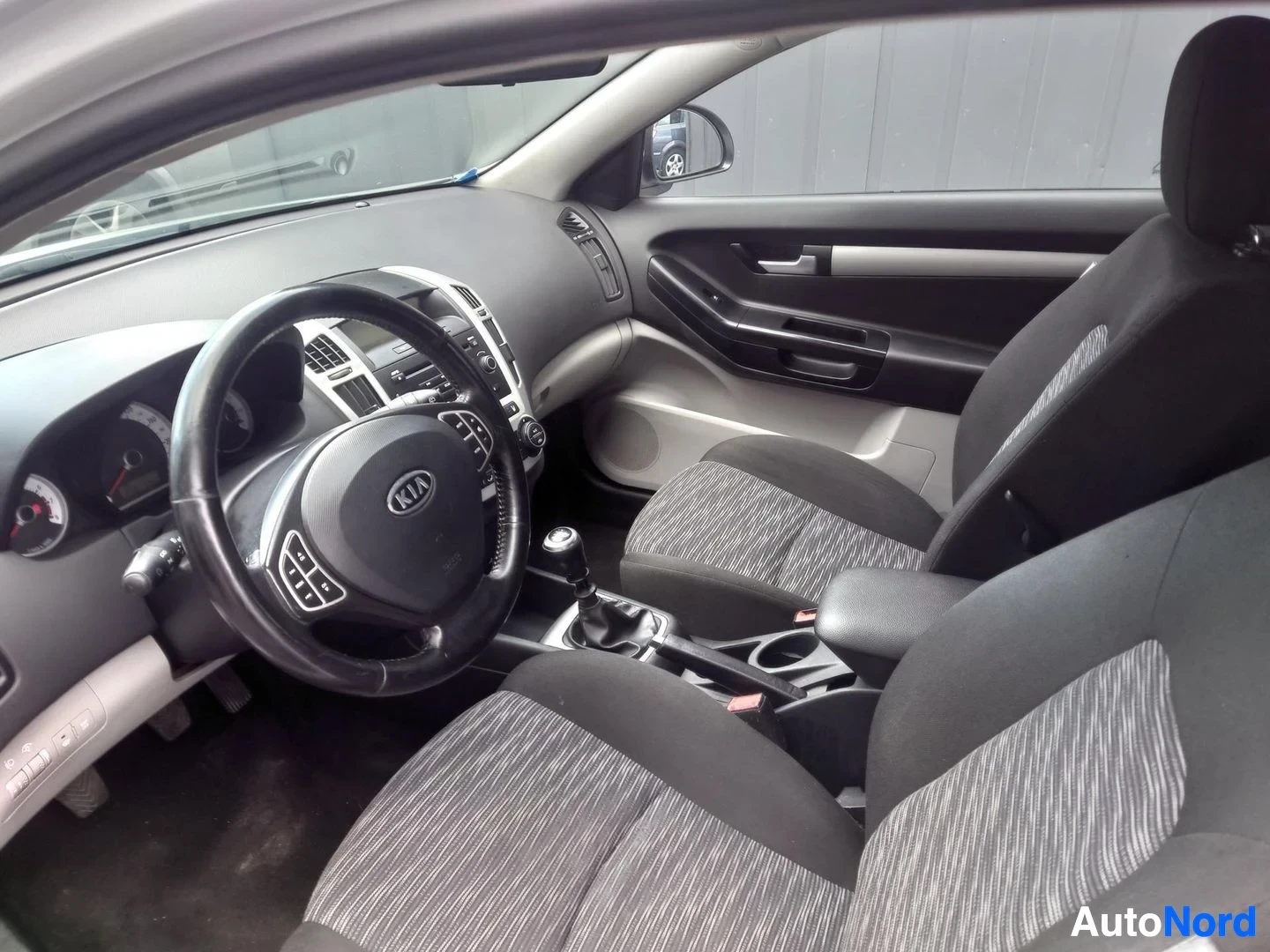 Muu: Kia ProCeed 2008 kuljettajan puolen sisätila: KIA-merkkinen ohjauspyörä, manuaalivaihteiston keppi ja tummat kankaanistuimet.