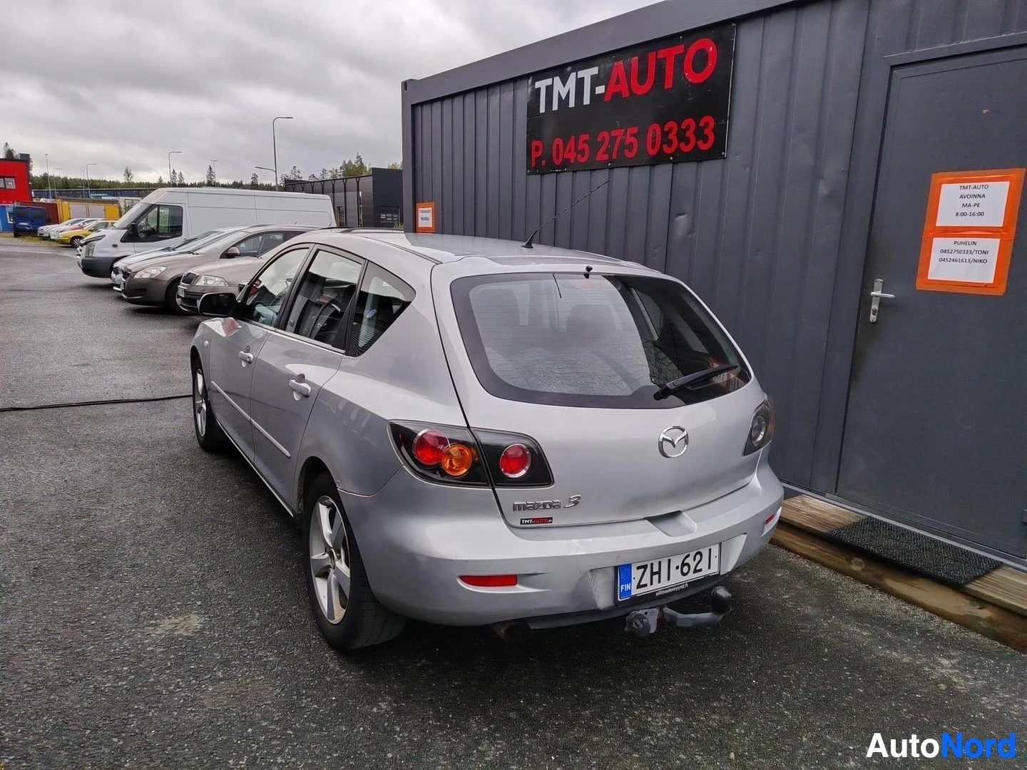 Takaviisto: Hopeinen Mazda 3 -viistoperä kuvattuna takaosalta ja vasemmalta sivulta; rekisterikilpi, perässä vetokoukku; taustalla TMT-AUTO-rakennus ja muita parkkeerattuja