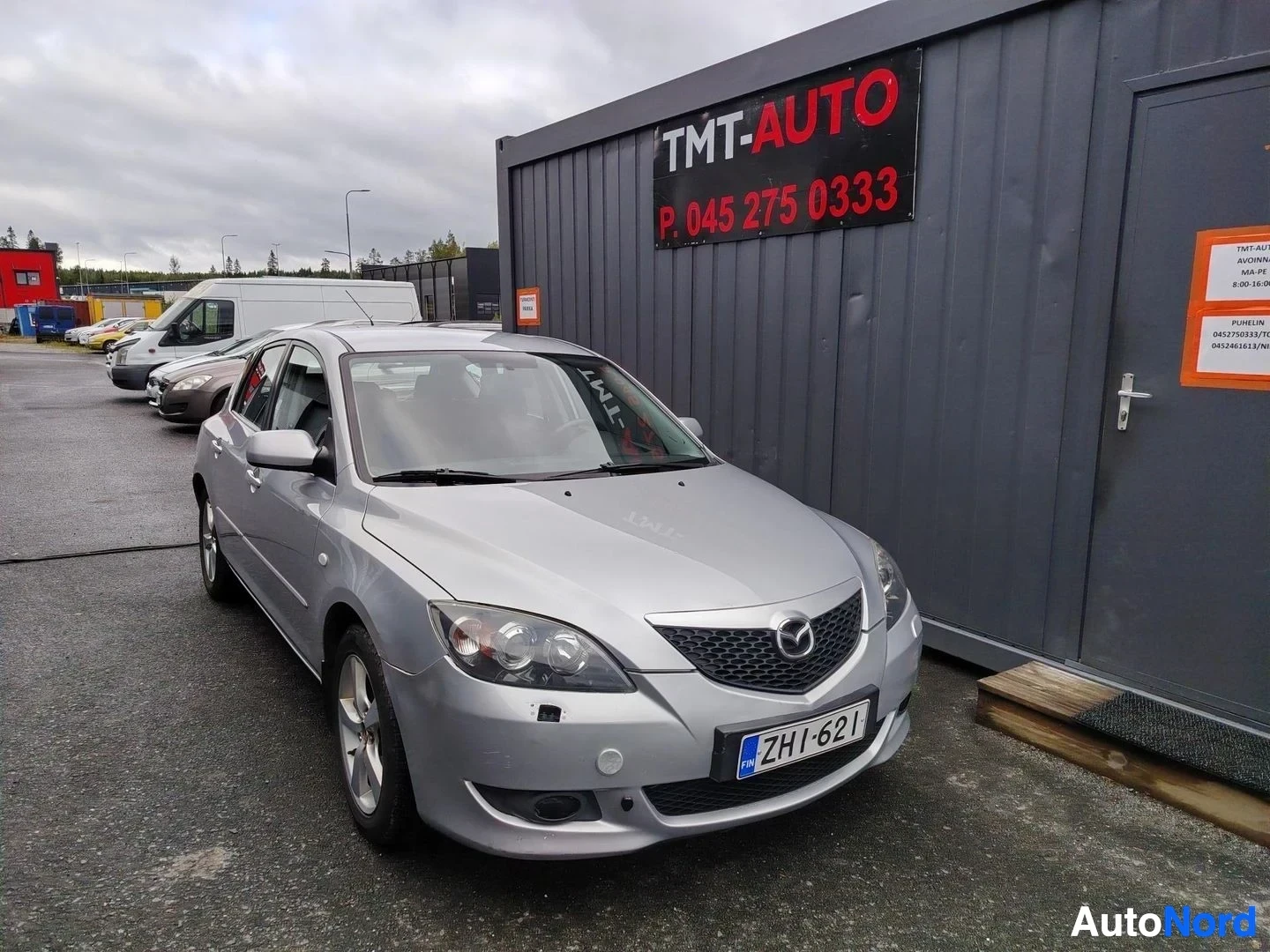 Etuviisto, nokka oikealle: Hopeanharmaa Mazda 3 -henkilöauto kuvattu etukulmasta autonumerolla.