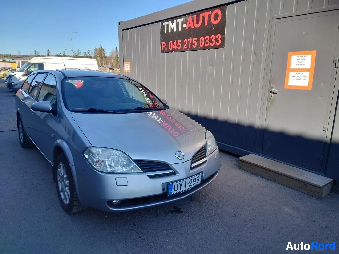 Etuviisto, nokka oikealle: Nissan Primera Farm, front three-quarter view, nose pointing to the right. Silver body, blue EU plate, parked beside a gray TMT-AUTO container building.