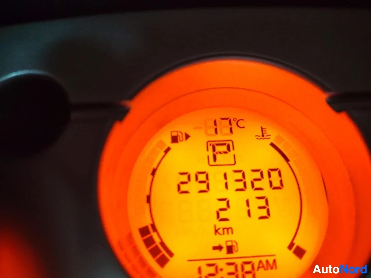 Yksityiskohta: Kojelautanäyttö oranssilla taustalla näyttää matkamittarin lukeman 291320 km ja ajan 12:38 AM.