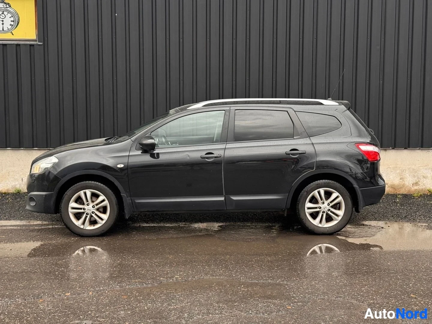 Etuviisto, nokka vasemmalle: Mustan Nissan Qashqai+2:n kyljensuuntainen kuva vasemmalta; kattokaide, hopeavanteet ja luukkulasta näkyvät tumman seinän edessä.