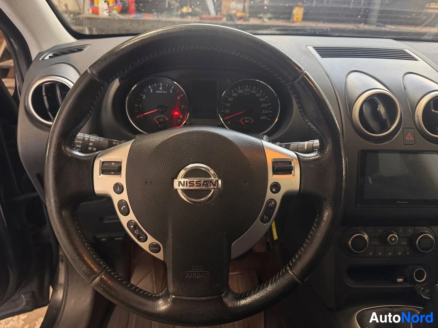 Muu: Ohjauspyörä lähikuvassa Nissan-merkillä, SRS AIRBAG-teksti, mittarit ja kojelaudan ilmastointilähetteet.