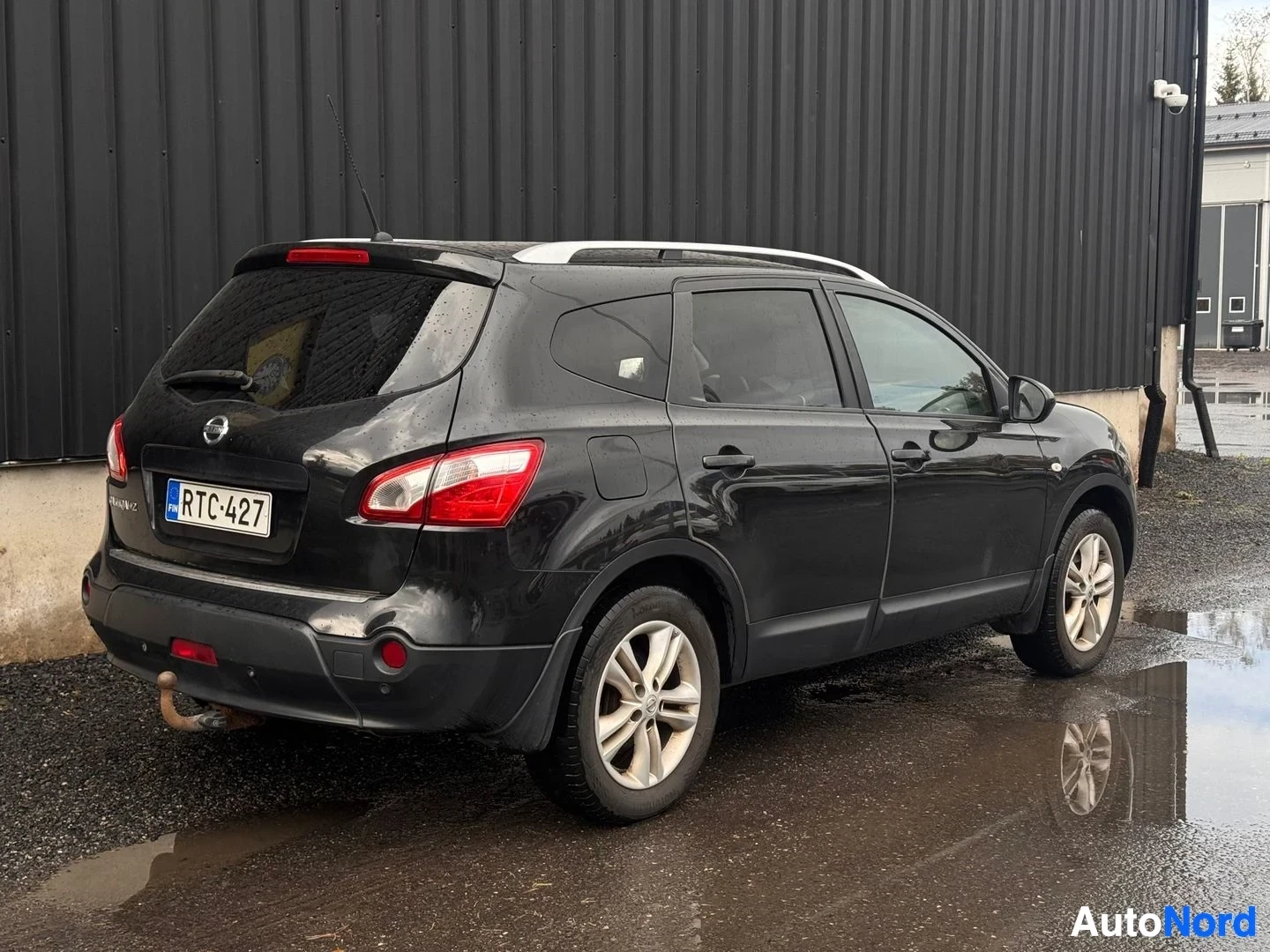 Takaviisto: Mustan Nissan Qashqai+2:n takakulmakuva vasemmalta; rekisterikilpi, takavetoiskukoukku, kattokaiteet ja hopeavanteet märällä pihalla.