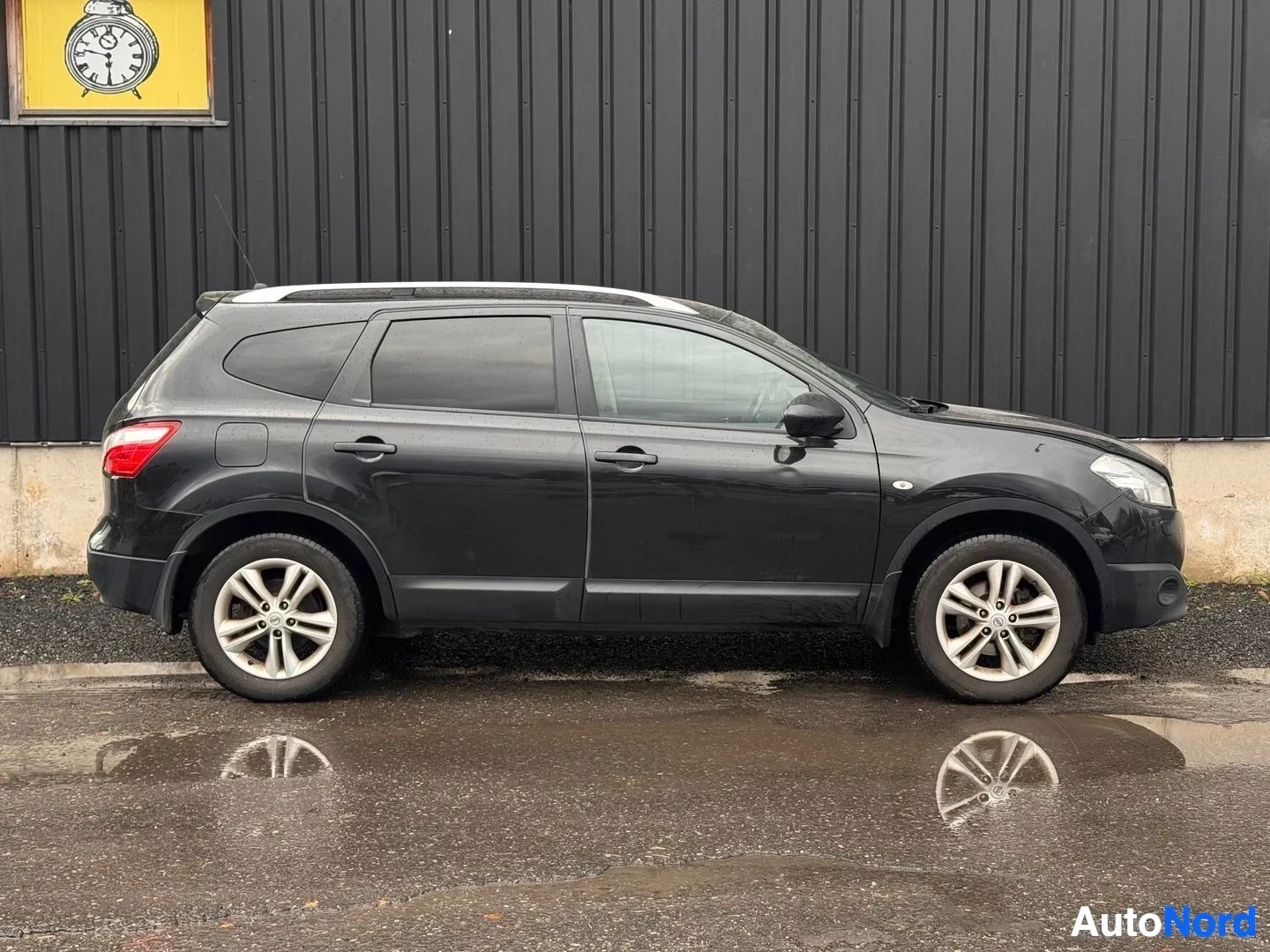 Takaviisto: Musta Nissan Qashqai +2 -farmarityylinen auto sivusta-takaa kuvattuna, kattokaiteet ja takavalot näkyvillä, märkä asfaltti alusta.