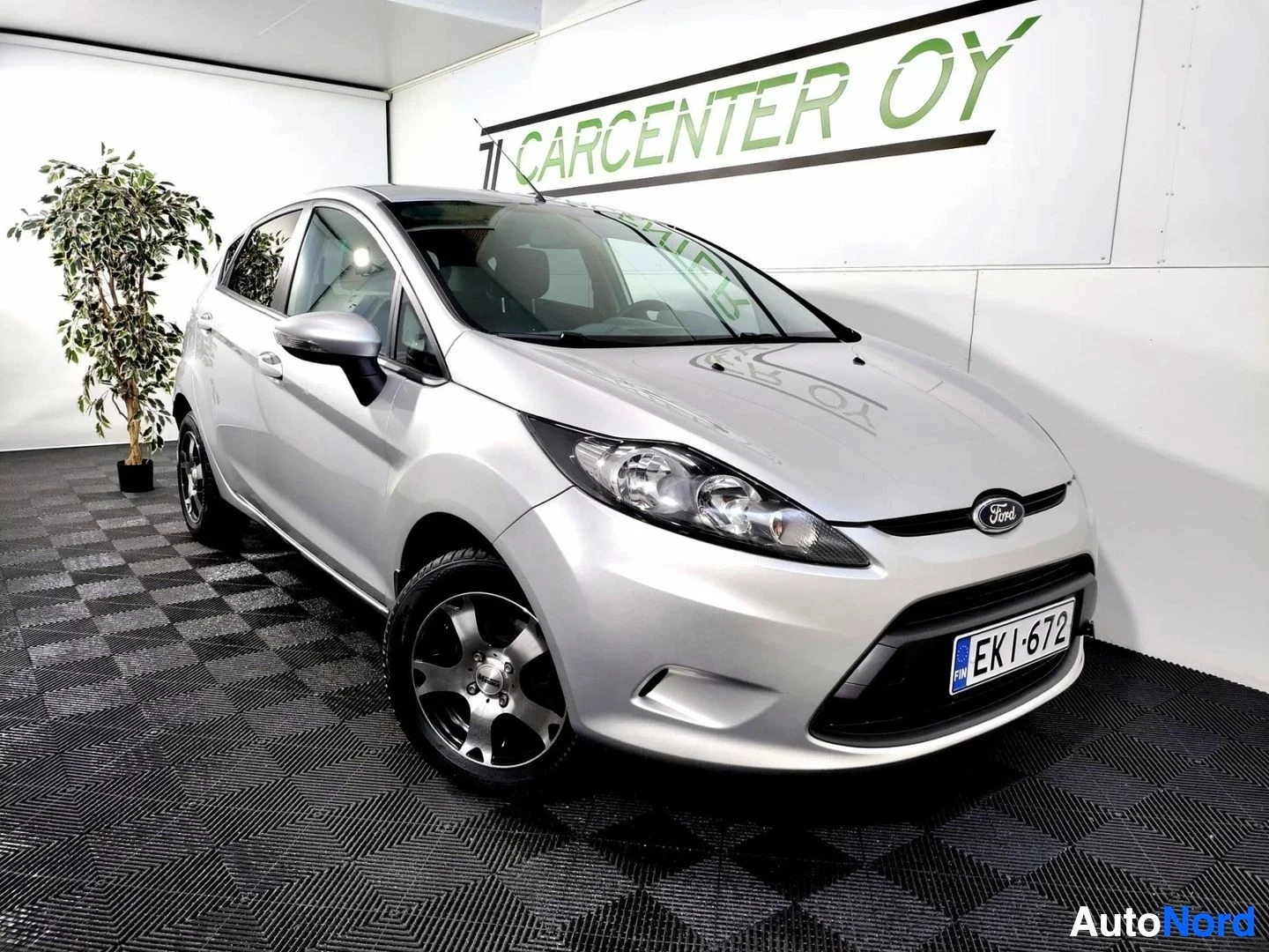 Etuviisto, nokka oikealle: Hopeanvärinen viisiovinen Ford Fiesta -henkilöauto etuviistosta kuvattuna autonäyttelytilassa, rekisterikilpi.