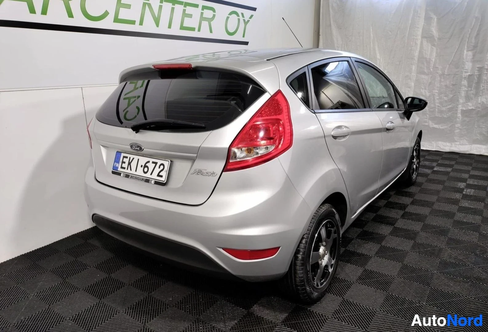Takaviisto: Hopeanvärinen Ford Fiesta -henkilöauto takaviistosta kuvattuna sisätiloissa, takapuskuri ja rekisterikilpi näkyvät.