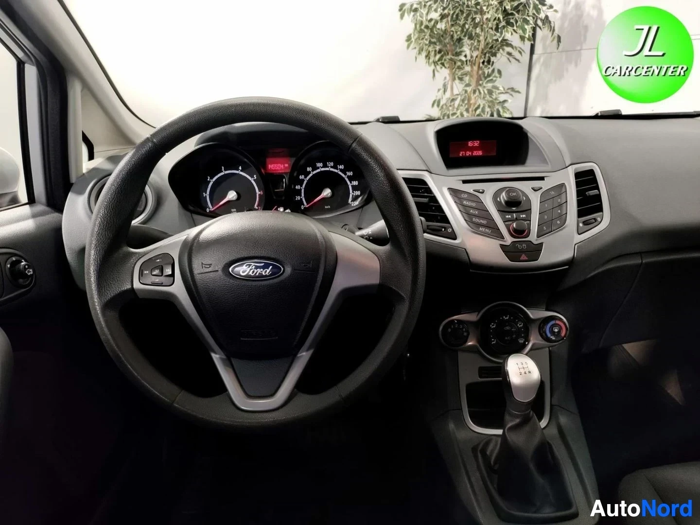 Edestä: Lähikuva ohjauspyörästä ja kojelaudasta: Ford-embleemi, mittaristo, keskikonsolin radio- ja ilmastointisäätimet sekä manuaalivaihteen keila.