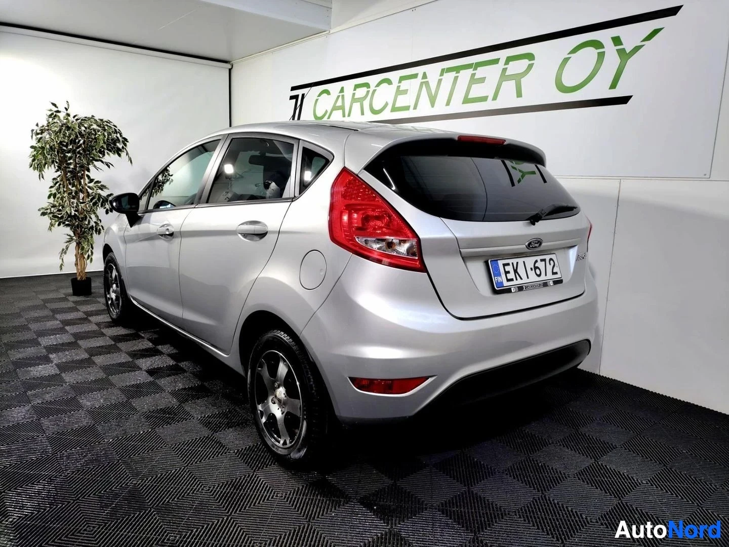 Takaviisto: Hopeanvärinen Ford Fiesta -henkilöauto takaviistosta kuvattuna sisätiloissa, takavalot ja rekisterikilpi näkyvät.