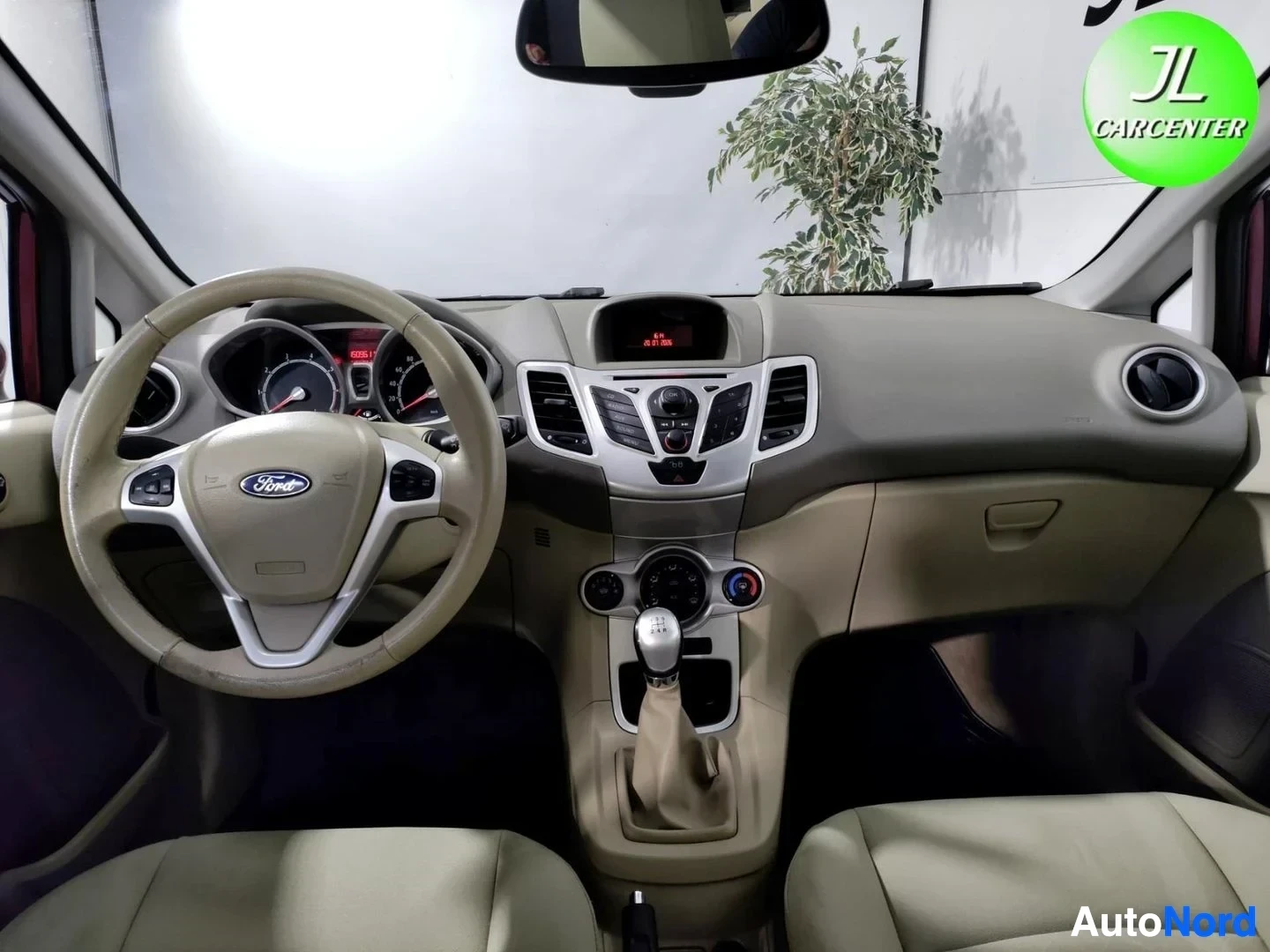 Muu: Näkymä ohjaamoon tuulilasin läpi: beige kojelauta, Ford-merkkinen ohjauspyörä, keskikonsoli ja manuaalivaihteen keilu.