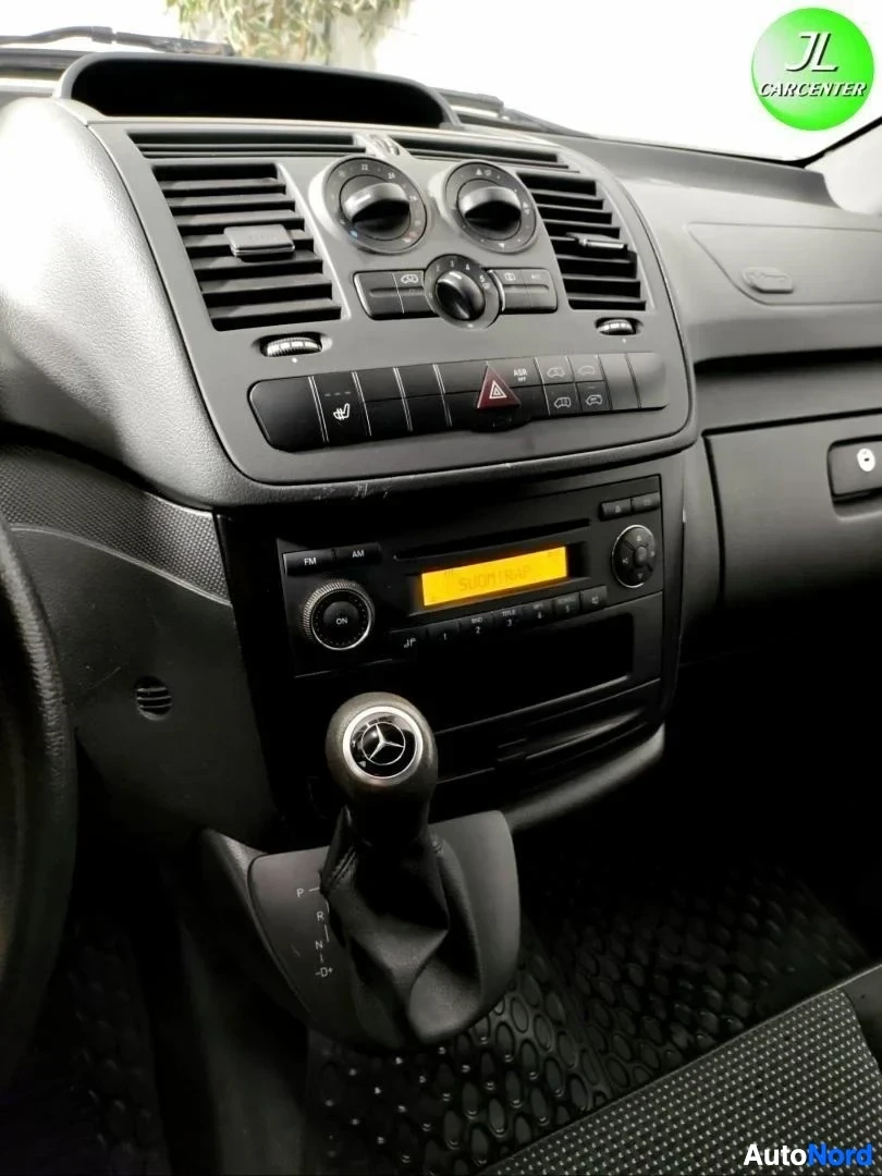Sivulta: Kojetaulun keskiosan lähikuva: ilmastointisäätimet, varoituskolmio ja radio CD-soittimen kanssa, alla Mercedes-merkitty vaihteenvalitsin.