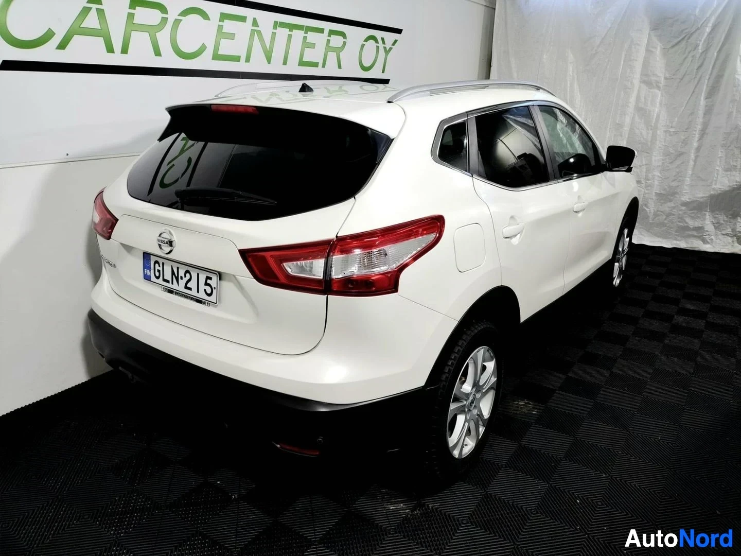 Takaviisto: Valkoisen Nissan Qashqain takakulmanäyttö: rekisterikilpi, kattokaiteet ja alloy-vanteet näkyvissä sisätiloissa.