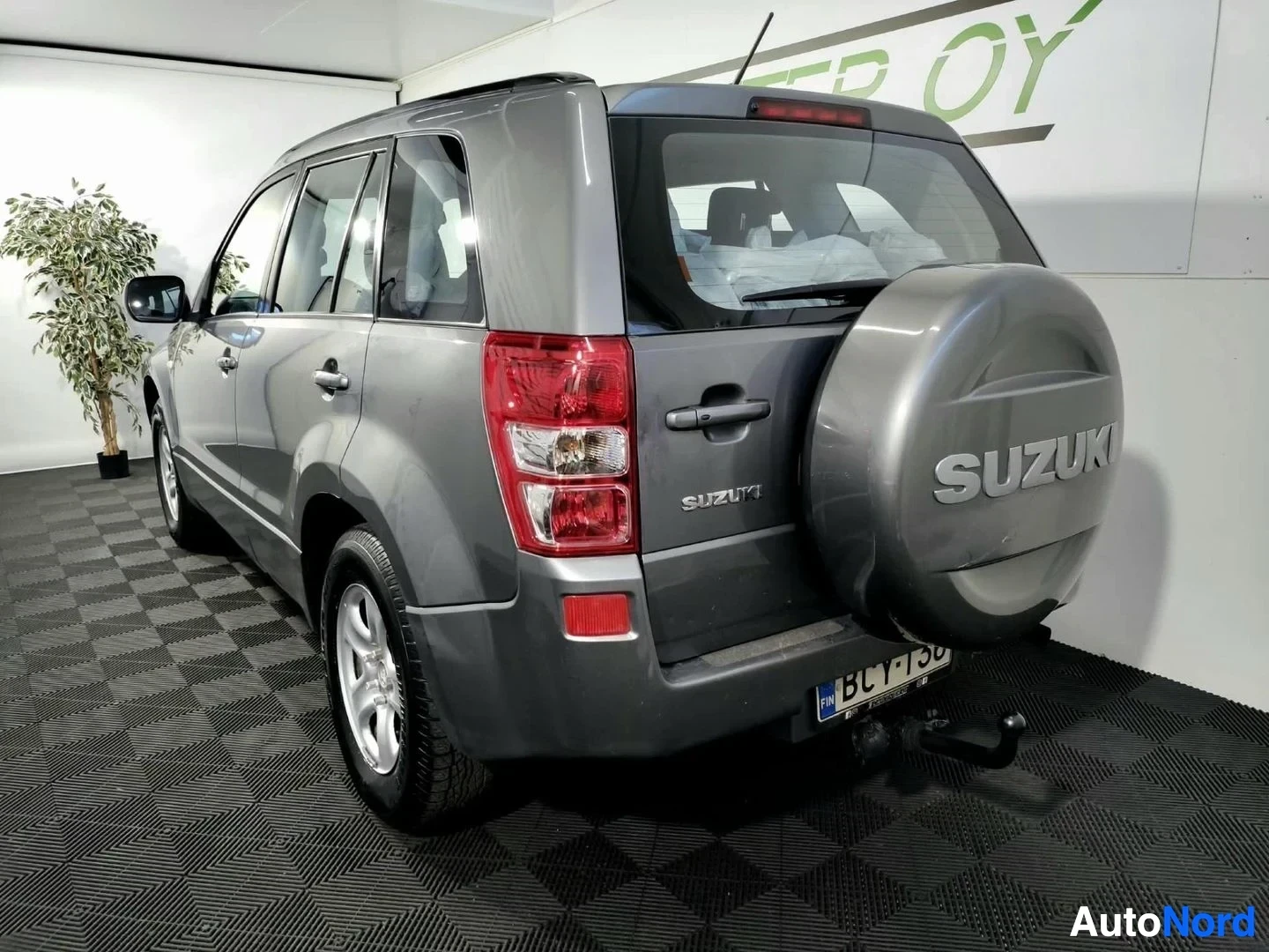 Takaviisto: Auton takaosa ja vasen sivu kolmen neljäsosan kulmasta; takavalot, varapyörä SUZUKI-tekstillä, rekisterikilpi ja kytketty vetokoukku harmaassa sisätilassa.