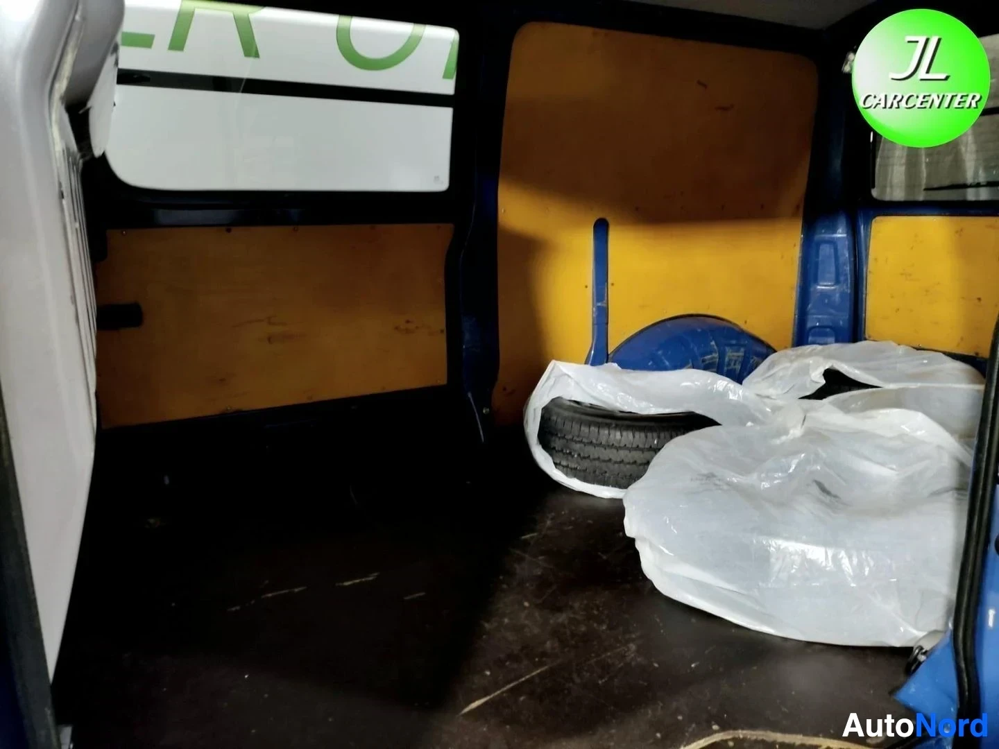 Tavaratila: Pakettiautan tavaratila: keltainen vaneripaneelit, lattialla renkaita muovipusseissa, tumma likainen lattia.