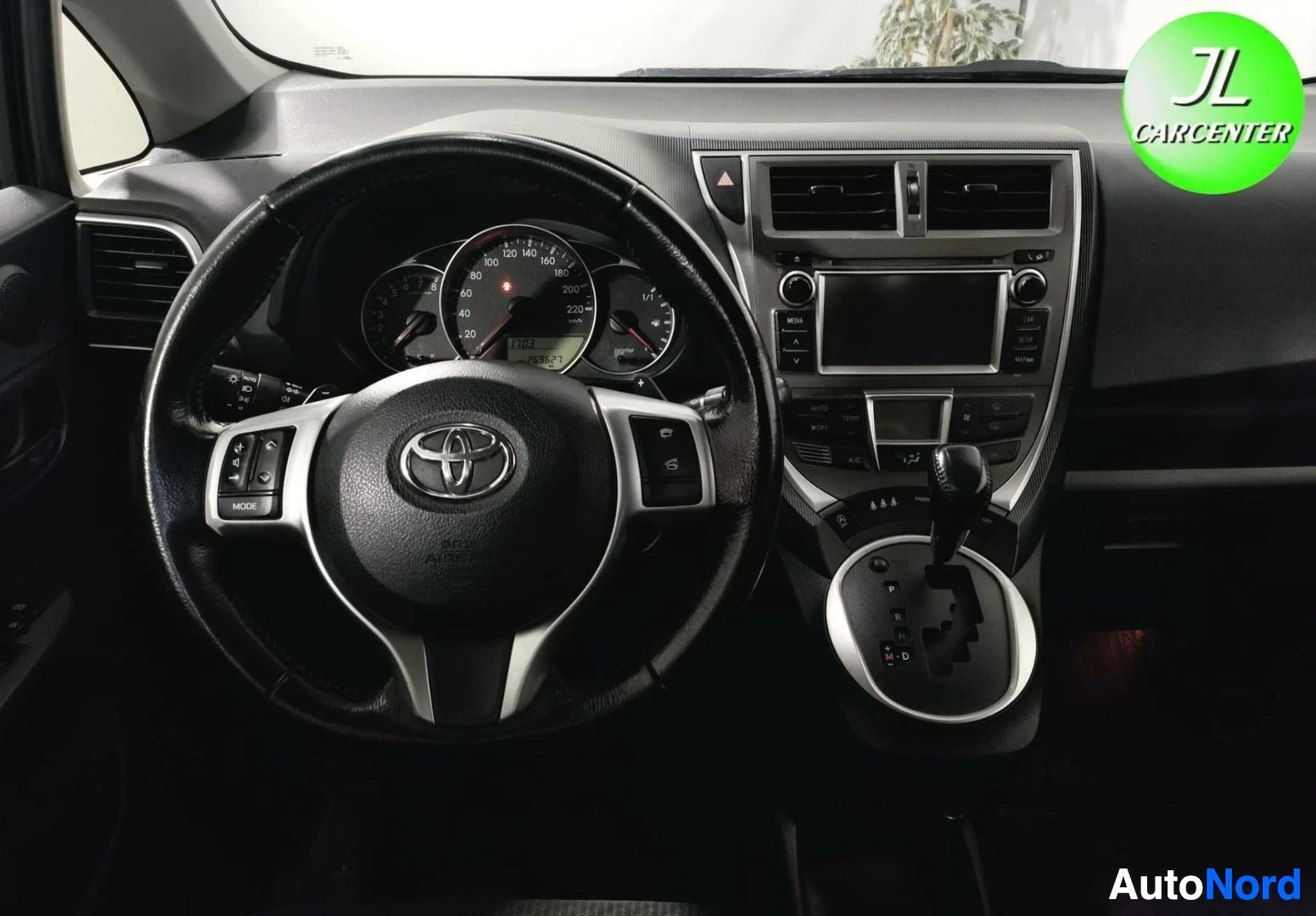 Muu: Ohjauspyörä ja kojelauta lähikuvassa: Toyota-merkki, mittariston ympyrämittarit, kosketusnäyttö ja automaattivaihteen valitsin.