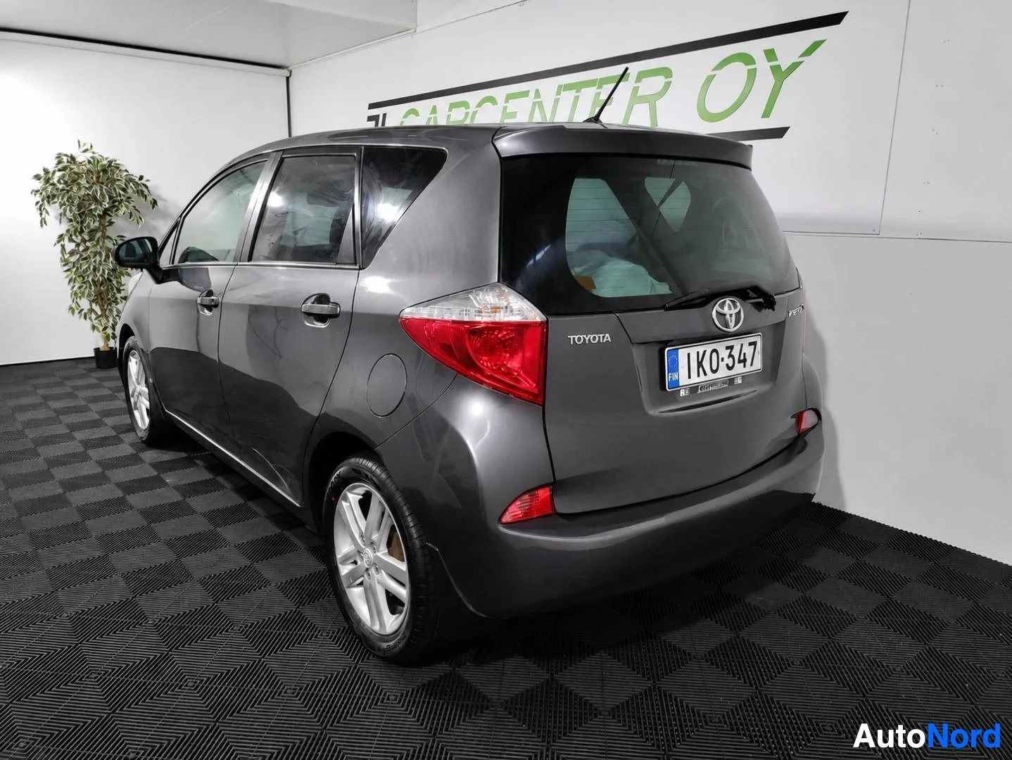 Takaviisto: Tummanharmaa Toyota Verso-S takaviistoon oikealta kuvattuna, takavalo ja TOYOTA-teksti näkyvissä.