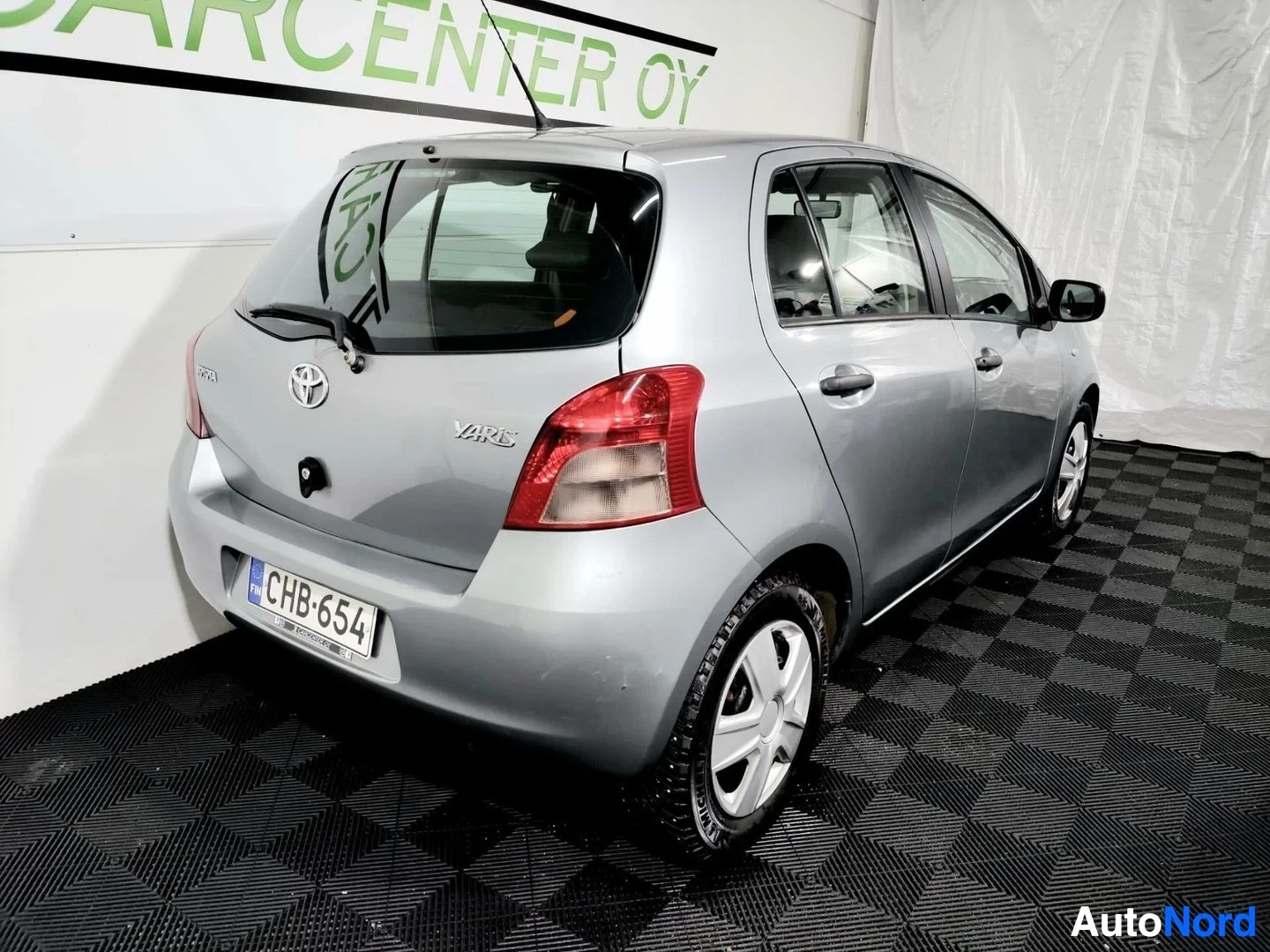 Takaviisto: Hopeanharmaa Toyota Yaris -viisiovinen takaa ja vasemmalta sivulta kuvattuna, rekisteritunnus.