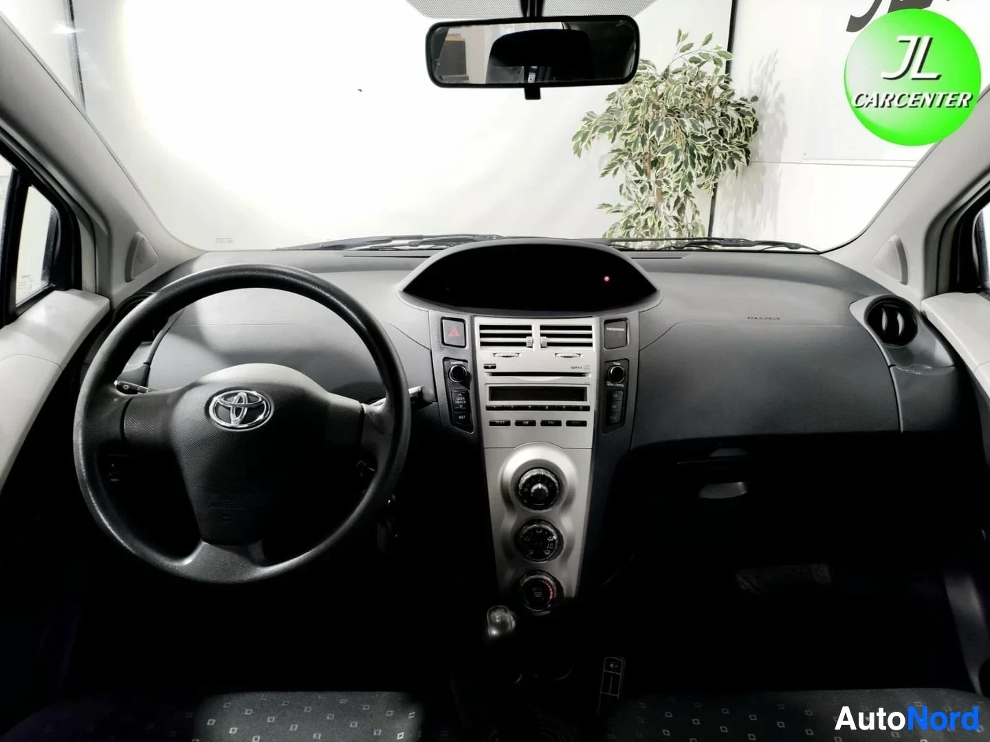 Muu: Ohjaamo etuosalta: Toyota-merkkinen ohjauspyörä, tumma kojelauta, keskikonsoli ja tuulilasin takana vihreä kasvi.