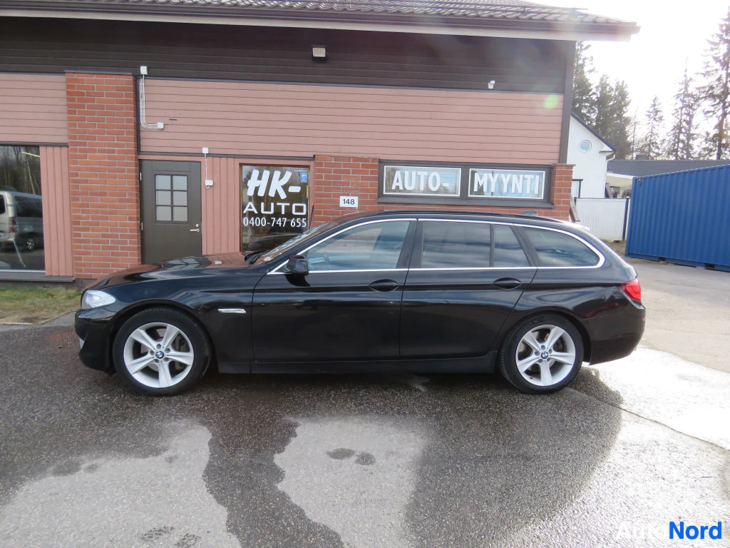Sivulta: Musta BMW 5-sarjan farmari sivulta, omakorkeus, hopeavanteet, autoliikkeen rakennuksen edessä.