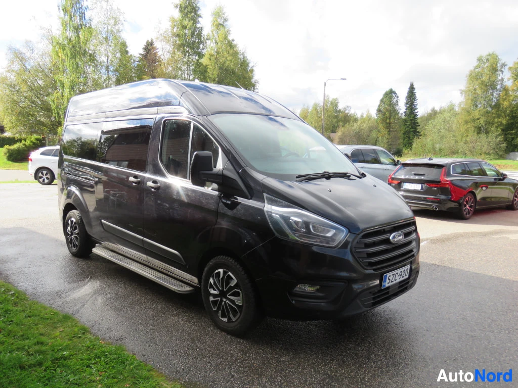 Etuviisto, nokka oikealle: Musta Ford Transit Custom -pakettiauto etuoikealta kuvattuna, koko auto näkyvissä, rekisterikilpi.