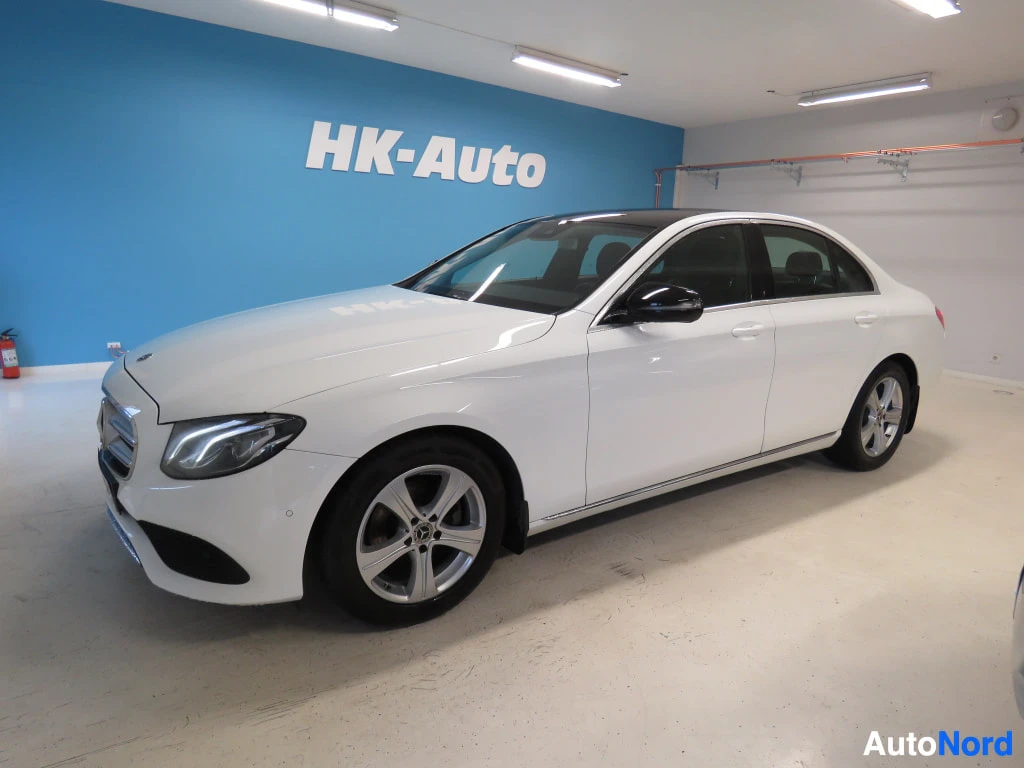 Etuviisto, nokka vasemmalle: Mercedes-Benz E-sarjan sedan etuviistosta vasemmalta, valkoinen kori ja musta lasikatto, sininen HK-Auto-taustaseinä.