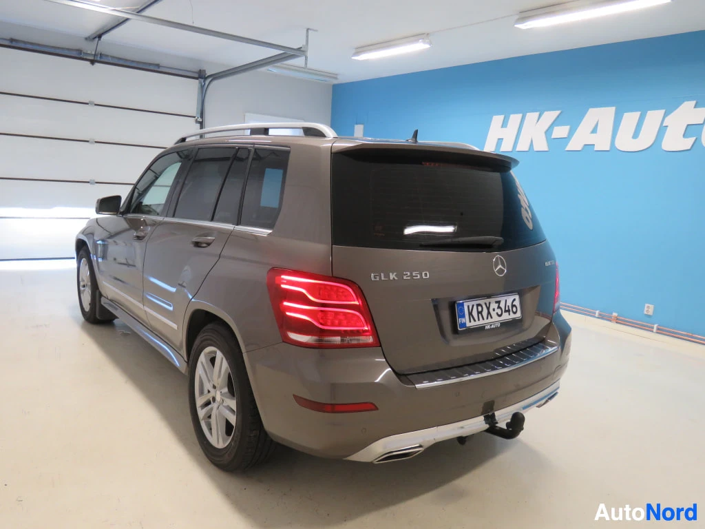 Takaviisto: Mercedes-Benz GLK:n takakulmanosto: takavalot, GLK 250 -merkintä, rekisterikilpi ja vetokoukku näkyvät sinistä HK-Auto-seinää vasten.